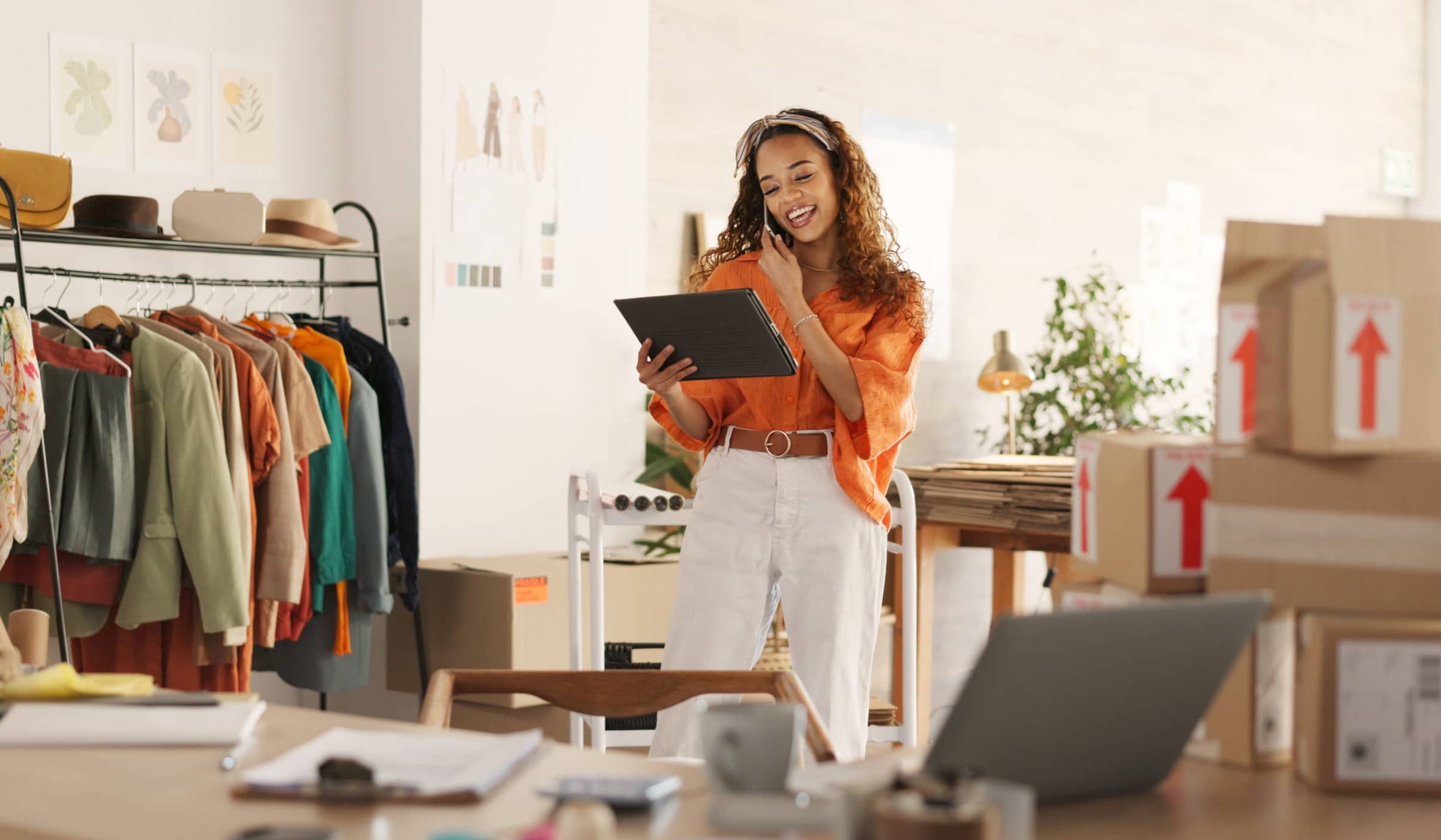 Smiling woman using a tablet and phone in a small business workspace surrounded by shipping boxes and clothing racks, representing Fexco’s international payment solutions for SMEs.